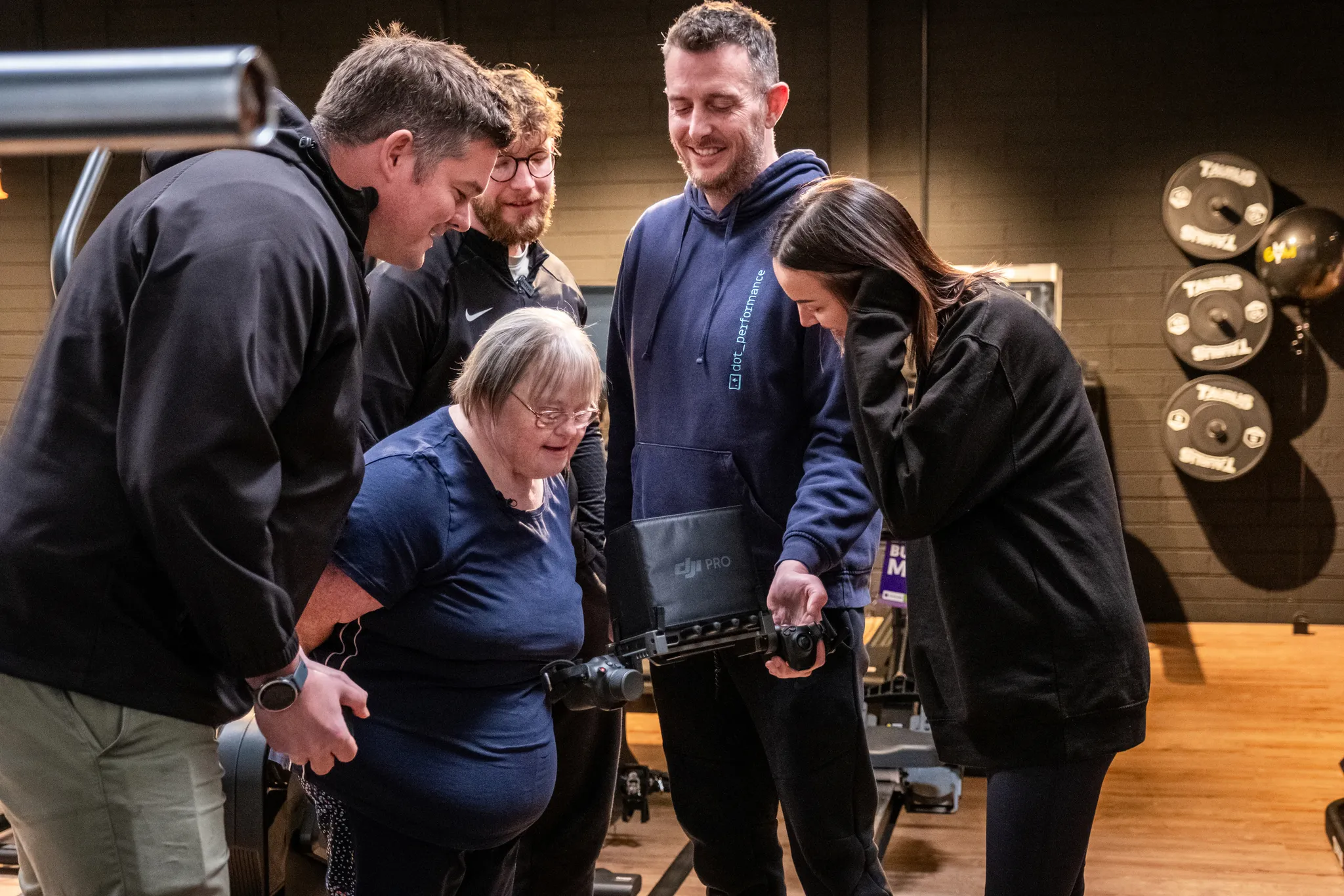 A group of four gym staff and trainers gathered around a woman with Down's syndrome in a fitness studio, looking at footage on a DJI Pro camera, with weight plates visible on the wall in the background